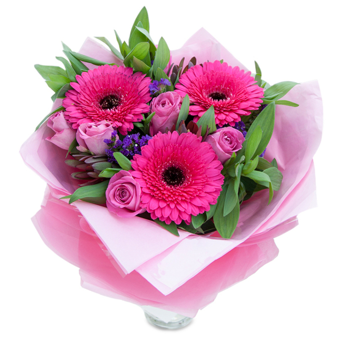 I often describe bouquets to customers as little stories; this one opens with three confident fuchsia Gerbera daisies that demand attention with their broad, radiating petals and dark brown-black centres that act like punctuation marks in the composition. Nestled closely among them are roses in a soft, dusty lavender-pink - each bloom still relatively tight, their velvet petals layered in a way that feels intimate and personal. A scattering of purple statice threads through the arrangement, introducing a papery, textured note and a cool counterpoint to the warm pinks. The greenery - think salal or eucalyptus - forms a generous, verdant backdrop: broad leaves with a slight sheen, a hint of early-morning dew, and the fresh scent of cut foliage. The bouquet is wrapped with a florist's care: an inner cradle of pale pink tissue paper supports the stems and softens the silhouette, while an outer veil of sheer fuchsia-pink cellophane, lightly crinkled, catches light and gives a gentle, celebratory sparkle. The lower portion shows the top rim of a clear glass vase, so the arrangement reads as both hand-tied and display-ready. The overall impression is one of playful grace - fragrant yet not overpowering - suited to marking a birthday, celebrating a mother, or offering congratulations. As a local florist familiar with Queen's Park, I imagine this piece greeting someone on a living room table near Gladstone Park or arriving as a bright surprise along Chamberlayne Road.